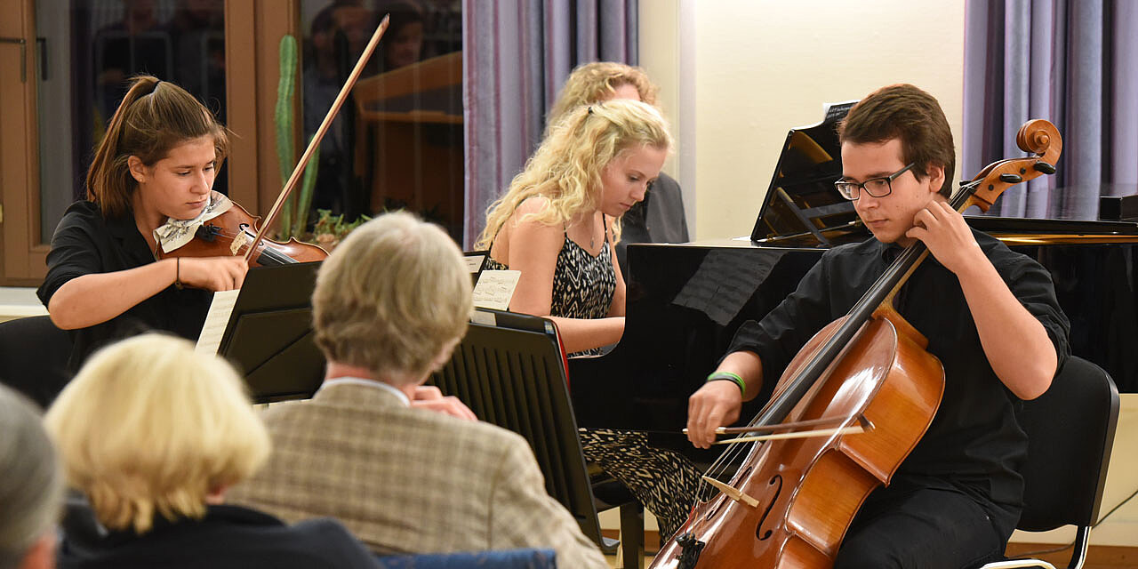 Im Vordergrund sieht man ein Publikum, welches drei Jugendlichen beim Geige, Cello und Flügel spielen zusieht.