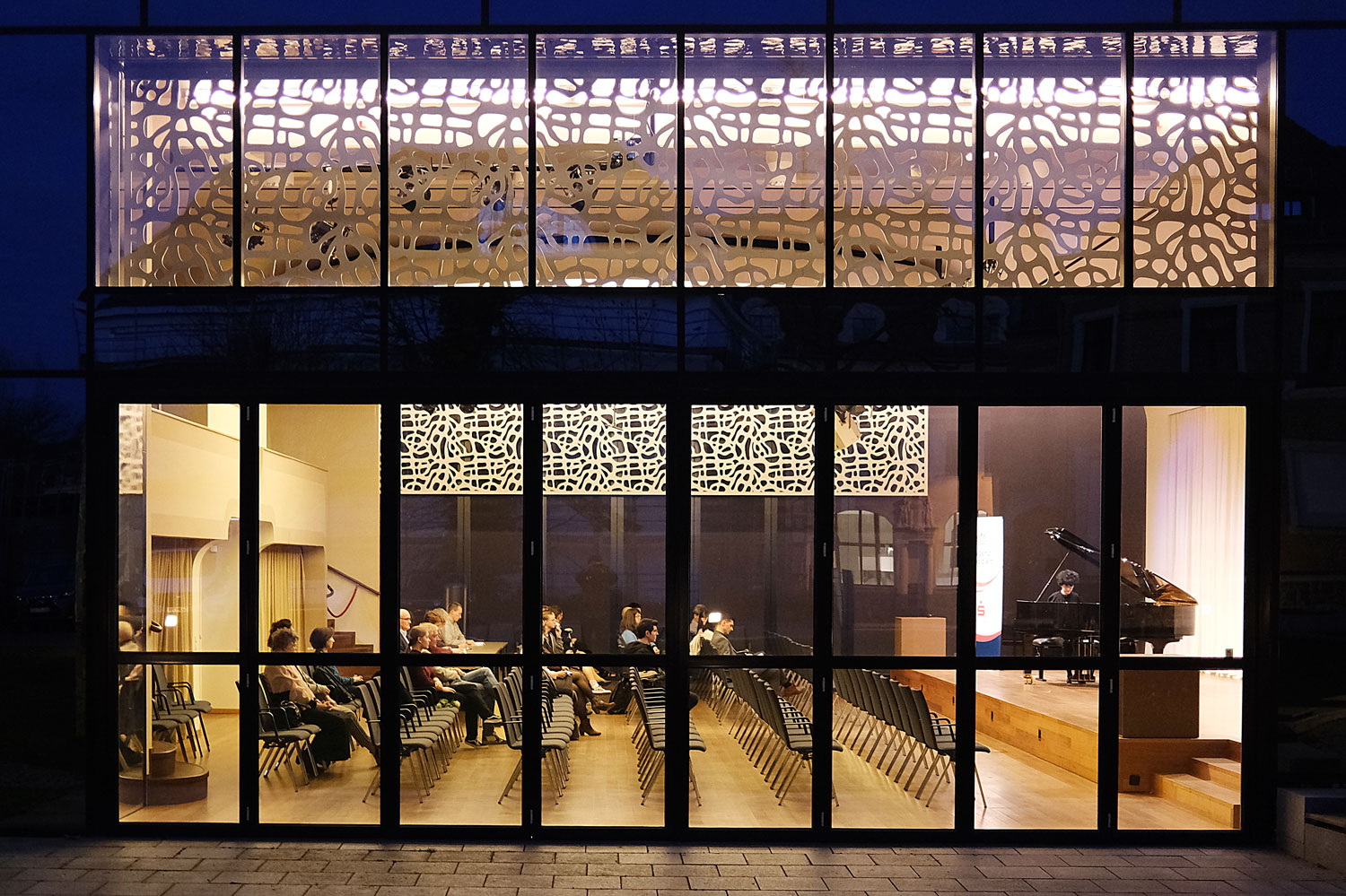 Blick von außen durch die Fensterfront des Konzertsaals in dem Publikum und rechts ein Pianist auf einer Bühne zu sehen ist