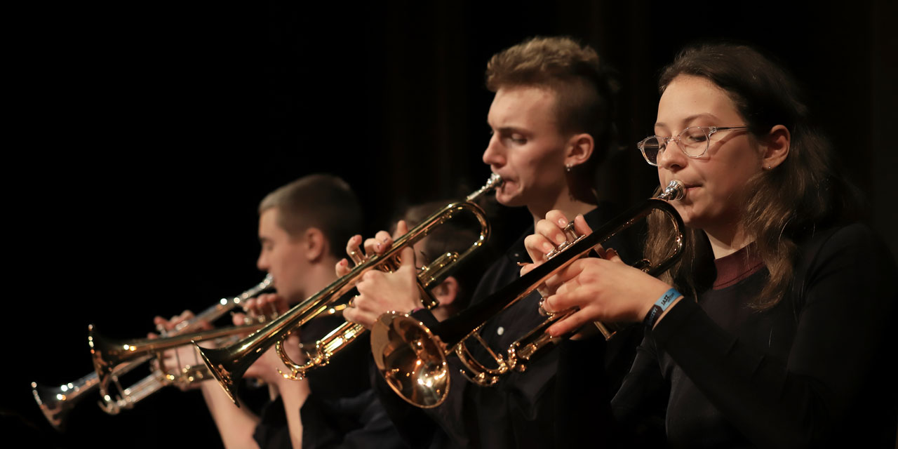 Vier schwarz gekleidete Personen spielen bei den Jazzwelten 2019Trompete. Eine von ihnen ist eine junge Frau mit braunen Haaren und Brille, ein andere hat blonde Haare in einem Irokesen-Schnitt 