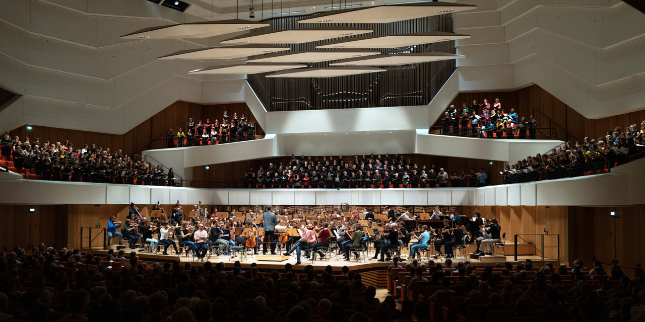 Auf der Bühne des Kulturpalast Dresdens spielt ein Sinfonieorchester, die Ränge hinter der Bühne sind mit mehreren Chören besetzt (aus dem Projekt Saxonia Cantat 2019) und auf der Bühne sitzen zwei Solisten Sänger-/innen, das Parkett des Saales ist mit Zuschauern gefüllt.