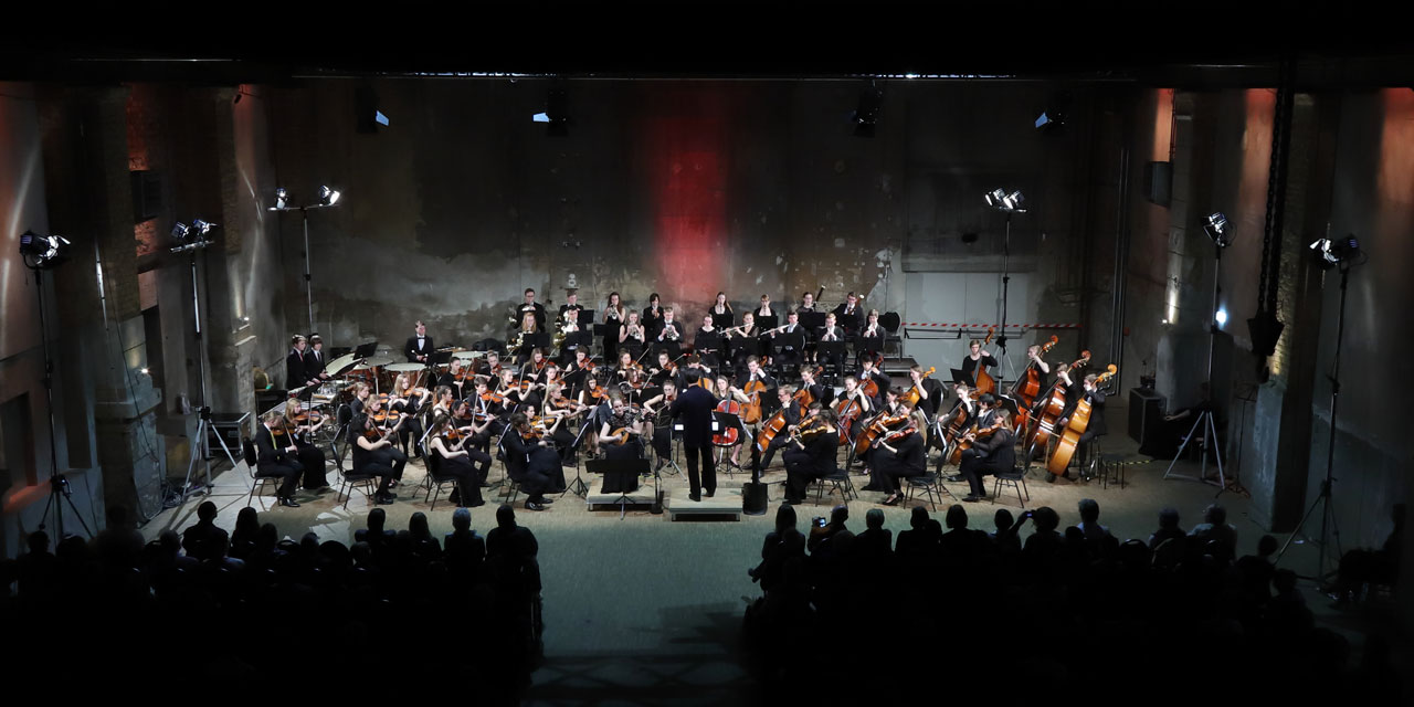 Das LJO Sachsen spielt in einer alten Industriehalle ein Konzert (2018), die Bühne ist hell beleuchtet und das Publikum bis auf den letzten Platz gefüllt.