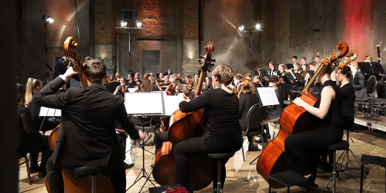 Im Fokus sind vier jugendliche Kontrabassspieler-/innen. Sie musizieren als Teil des LJO Sachsens 2018 in einer, dem Anschein nach älteren, ehemaligen Industriehalle. Alle Musiker-/innen sind schwarz gekleidet, es findet ein Konzert statt.