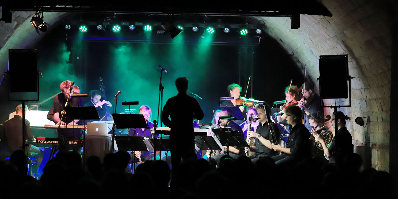 Das JJO Sachsen spielt in einem relativ niedrigen Gewölbe, die Bühne ist türkis beleuchtet und es liegt „Nebel“ in der Luft. Man sieht unter anderem Fagotte, Geigen und ein Klavier spielen.