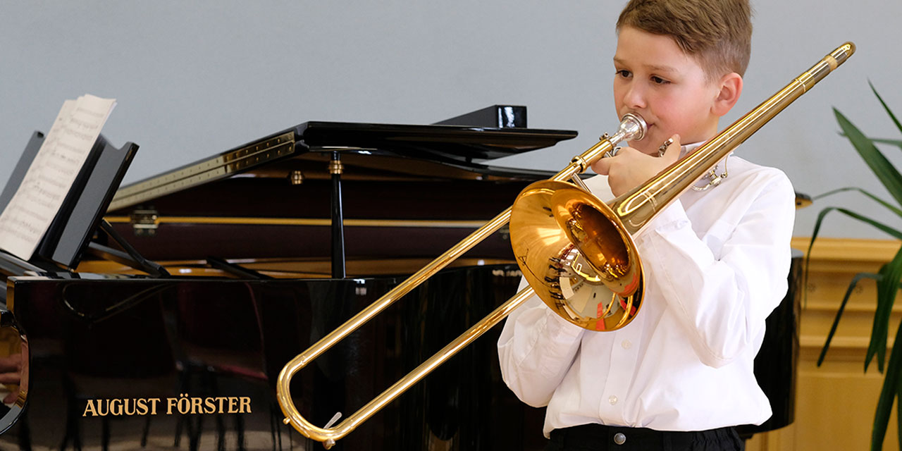 Ein Junge mit kurzen blonden Haaren, weißem Hemd und schwarzer Hose spielt beim Wettbewerb Jugend Musiziert Posaune. Im Hintergrund sieht man einen schwarzen Flügel, auf dem der Junge begleitet wird.