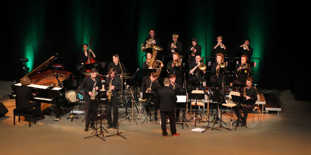 Das JJO Sachsen spielt auf einer Bühne vor schwarzem, grün angestrahltem Hintergrund. Alle Personen sind schwarz gekleidet, es sind Saxophone, Trompeten, Posaunen, Hörner, Klavier, Kontrabass und Schlagzeug vertreten.