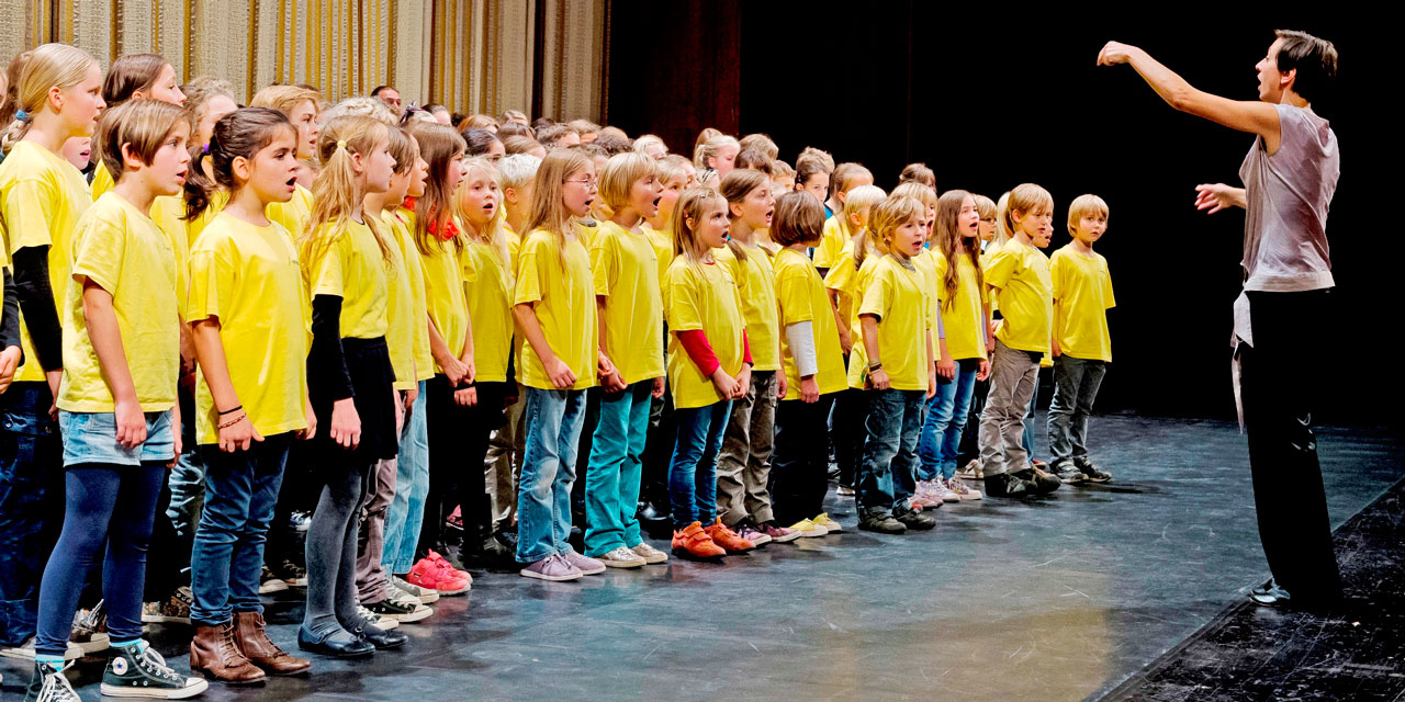 Die Dirigentin Sophie Bauer hebt die Hand zum Einsatz, öffnet den Mund und dirigiert einen Kinderchor. Die Kinder tragen gelbe T-Shirts und sind im Grundschulalter.