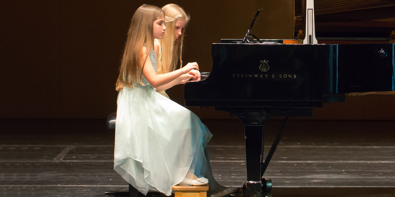 Zwei junge Mädchen mit langen hellen Haaren und hellblauen Kleidern sitzen an einem Flügel und spielen konzentriert gemeinsam das Instrument.
