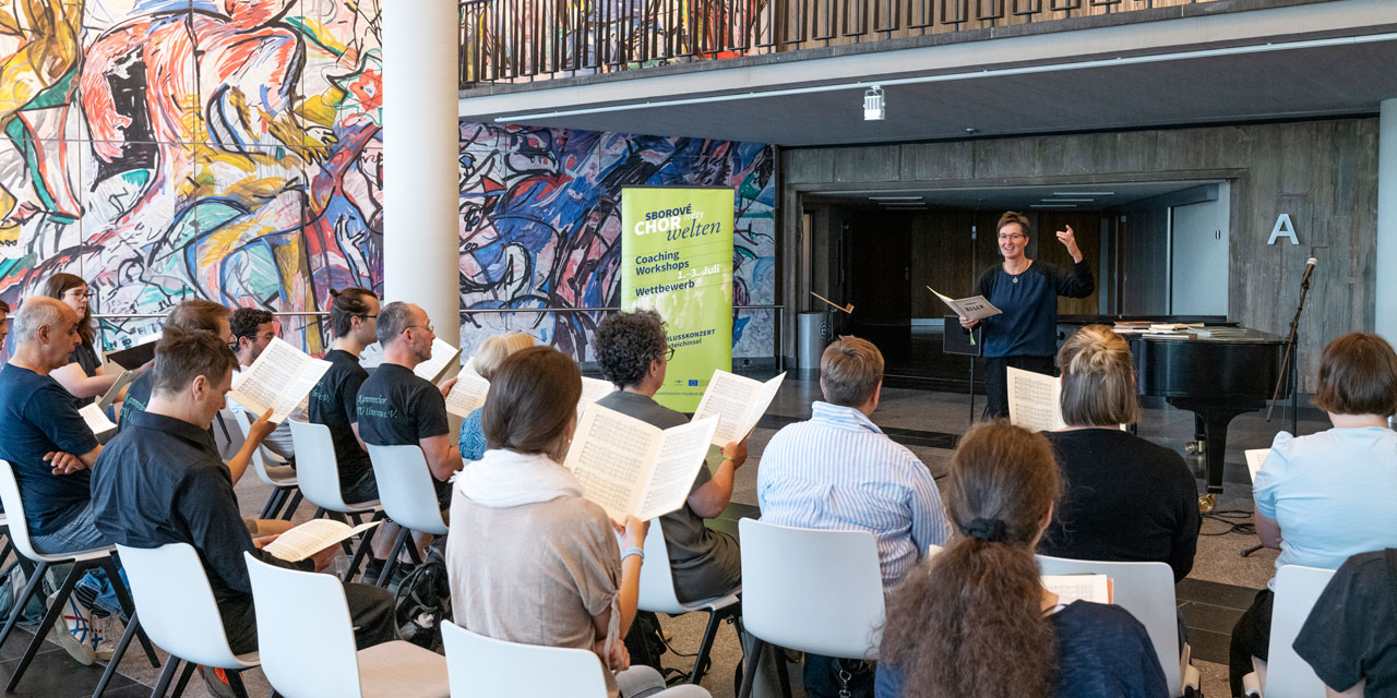 Eine Dirigentin leitet einen auf weißen Stühlen sitzenden Chor an