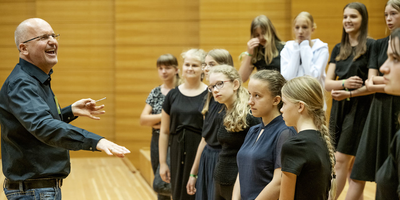 Ein Dirigent steht lachend vor einem Mädchenchor