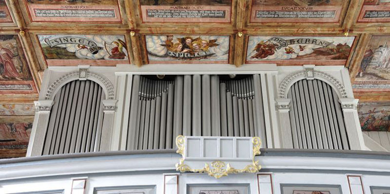 Hinter einer weißen, hölzernen Reling steht die ebenfalls weiße Orgel. Es sind lediglich drei „Fächer“ mit Pfeifen zu erkennen, die Orgel steht direkt unter bunt mit Bildern und