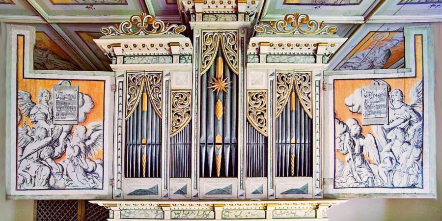 Es ist die Renaissance-Orgel der Wehrkirche Pomßen zu sehen. Auffällig ist die bunte Bemalung auf Orgel und Decke der Kirche. In fünf „Fächern“ kann man silberne, teils golden verzierte Orgelpfeifen erkennen, die größten in der Mitte, links und rechts gefolgt von den kleinsten und je noch weiter außen den Pfeifen mittlerer Größe. Oben an der Orgel und an manchen Pfeifen sind gelbgoldene Verzierungen angebracht. Die zwei aufgeklappten Schwingtüren der Orgel sind mit Bildern von Engeln, welche Noten halten, bestückt. 