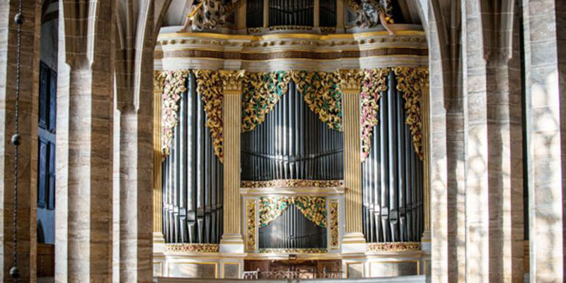 Die Silbermann-Orgel im Freiberger Dom ist in einiger Entfernung zwischen zwei Säulenreihen zu sehen. Gold-grüne bzw. gold-rote Verzierungen rahmen die silbernen Orgelpfeifen wie Vorhänge ein.