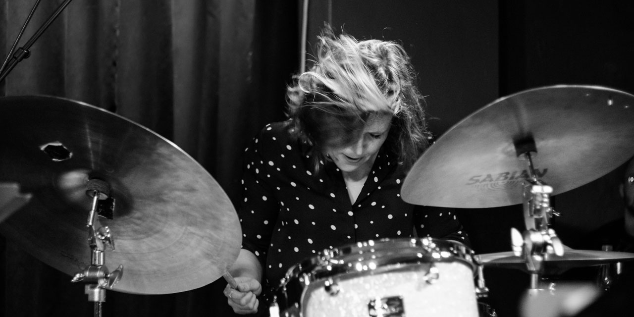 Eva Klesse, Schwarzweißfoto, Frau mittleren Alters, dunkles Oberteil mit hellen Punkten, Harre durch Kopfbewegung verweht, spielt Drumset, schlägt mit rechtem Stick Becken.