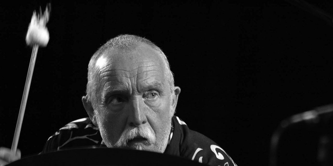 Günter Baby Sommer, Schwarzweißfoto, älterer Herr in Nahaufnahme, sehr kurze Haare, Bart, schwingt in rechter Hand einen Schlägel.