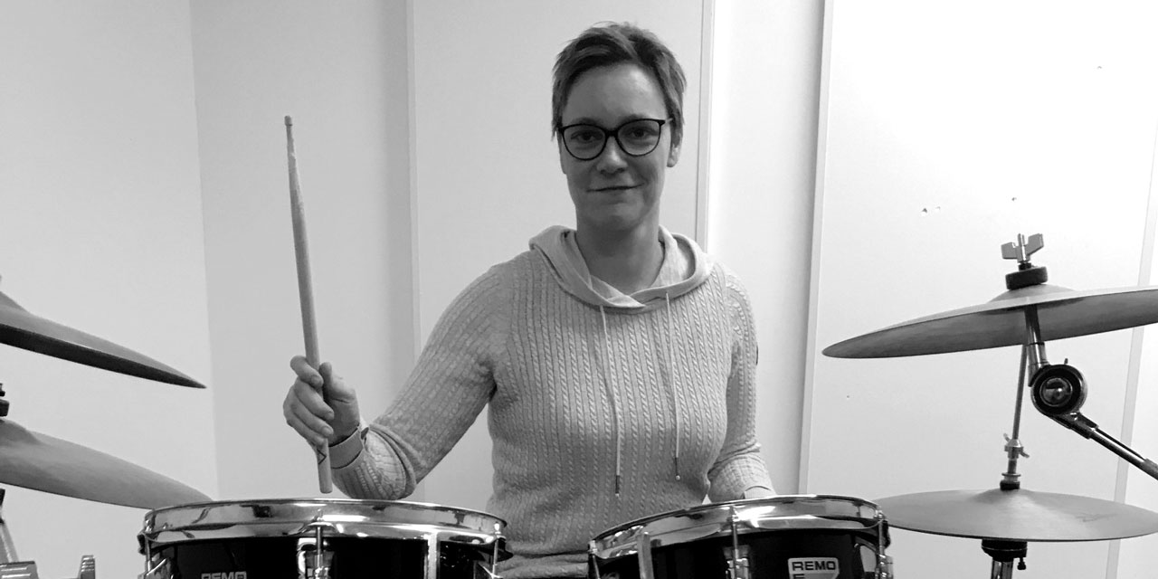 Nadine Berlt, Schwarzweißfoto, kurze Haare, Brille, sitzt vor Drumset und hält rechten Stick erhoben.