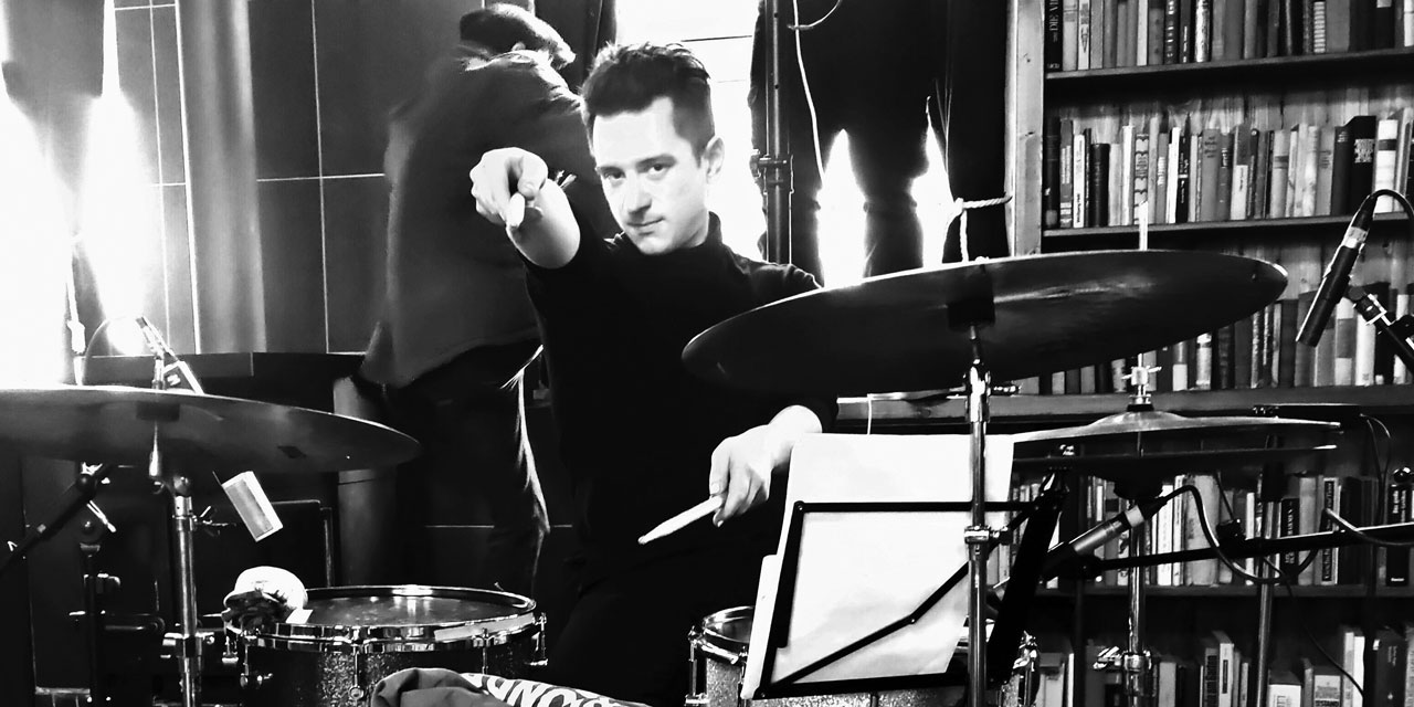 Philipp Scholz, Schwarzweißfoto, schwarzer Rollkragenpullover, sitzt vor Schlagzeug und zeigt mit rechtem Stick auf die Kamera, vor ihm steht Notenständer und auch ein Mikrofon ist links von ihm angebracht.
