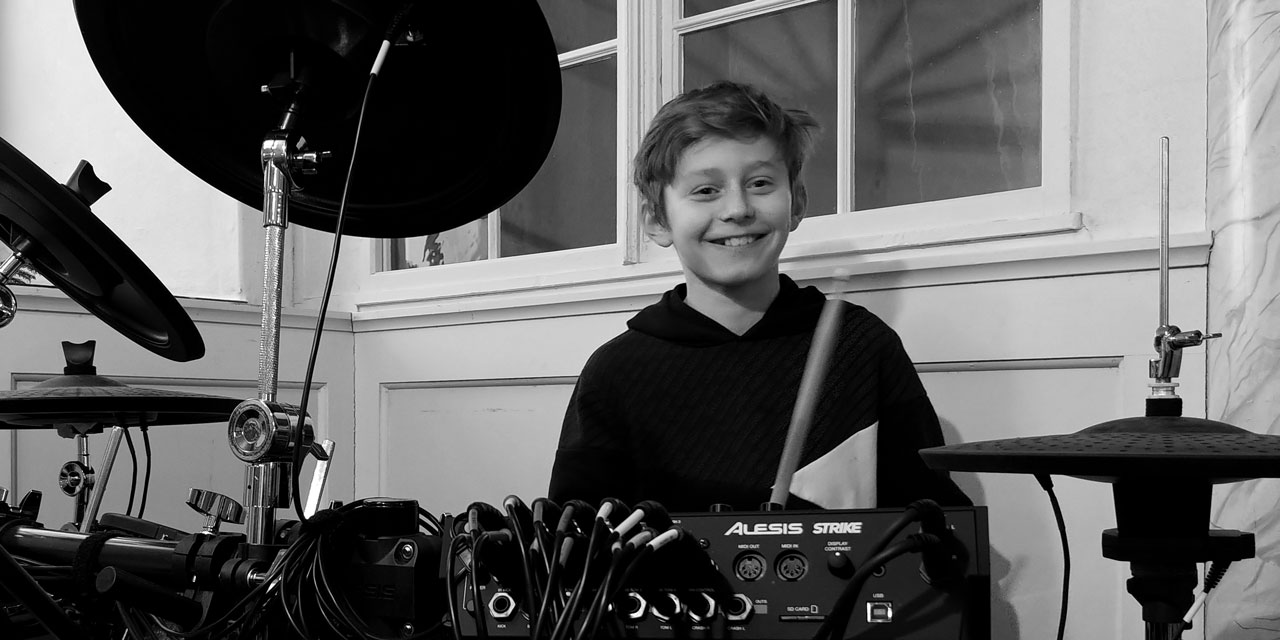 Tobias Trommler, Schwarzweißfoto, junger Teenager (13 Jahre), lächelt in die Kamera, sitzt hinter Mischpult mit (elektronischem) Drumset-Elementen, es sind 4 Becken zu sehen.