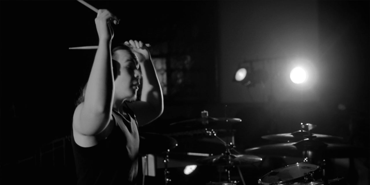Valentin Wutzler, Schwarzweißfoto, lange Haare, Kopfhörer auf, von Seite fotografiert, sitzt an Drumset und hat beide Arme mit Sticks erhoben.