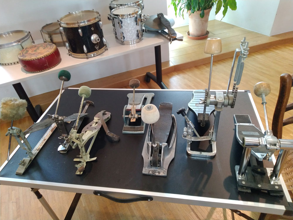 Auf einem Tisch steht eine Ansammlung an Pedalen, sogenannten „Fußmaschinen“, mit daran befestigten Schlägeln aus unterschiedlichen Materialien (bspw. Filz und Kunststoff). Im Hintergrund ist noch ein weiterer Tisch mit Trommeln verschiedener Größe, Farbe und Form zu erkennen.