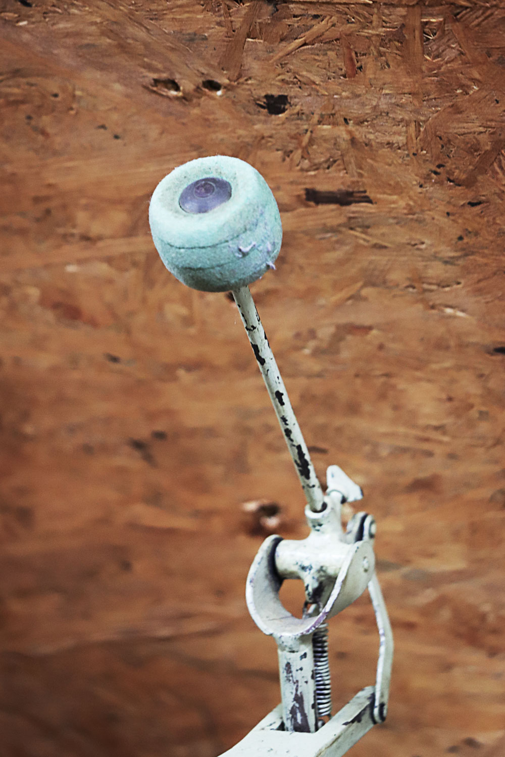 Ein Close-Up von einer Fußmaschine samt Filzschlägel vor einem Hölzernen Hintergrund. Der leicht bläuliche Lack der Konstruktion blättert schon an verschiedenen Stellen ab.