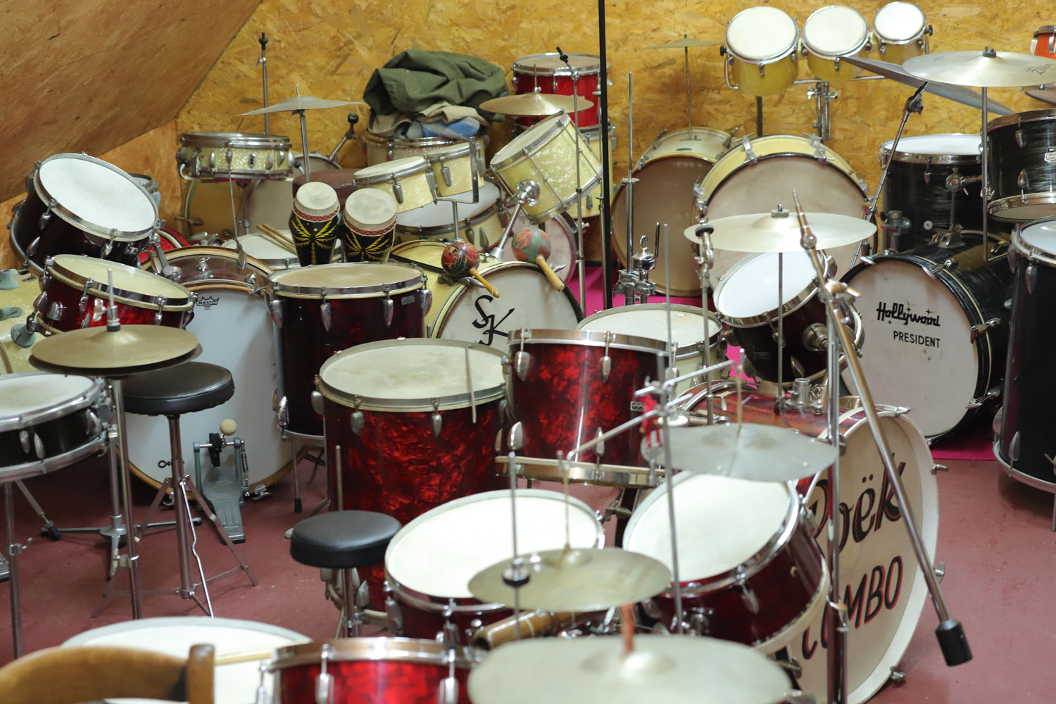 Man sieht eine große Sammlung verschiedenfarbiger Drumsets mit den verschiedensten Komponenten (z.B. Bongos, Becken, Tom’s und Base-Drums). Im Hintergrund ist die hölzerne Dachschräge des Raumes zu erkennen.