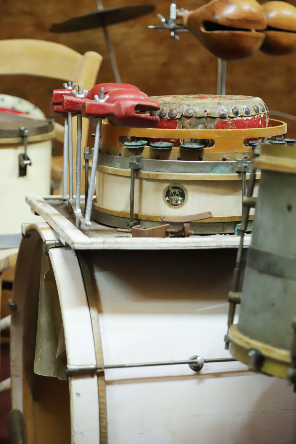 Eine Ansammlung mehrerer Schlagwerkobjekte ist übereinandergestapelt. Ganz unten steht eine gewaltige Trommel, bei der das Fell „zur Seite“ zeigt. Darauf befinden sich auf einer Holzunterlage zwei kleinere Trommeln mit horizontalem Fell, ein Schelleninstrument und drei rote, nebeneinander an vertikalen Metallstäben angebrachte, krabbenscherenförmige Klangkörper.