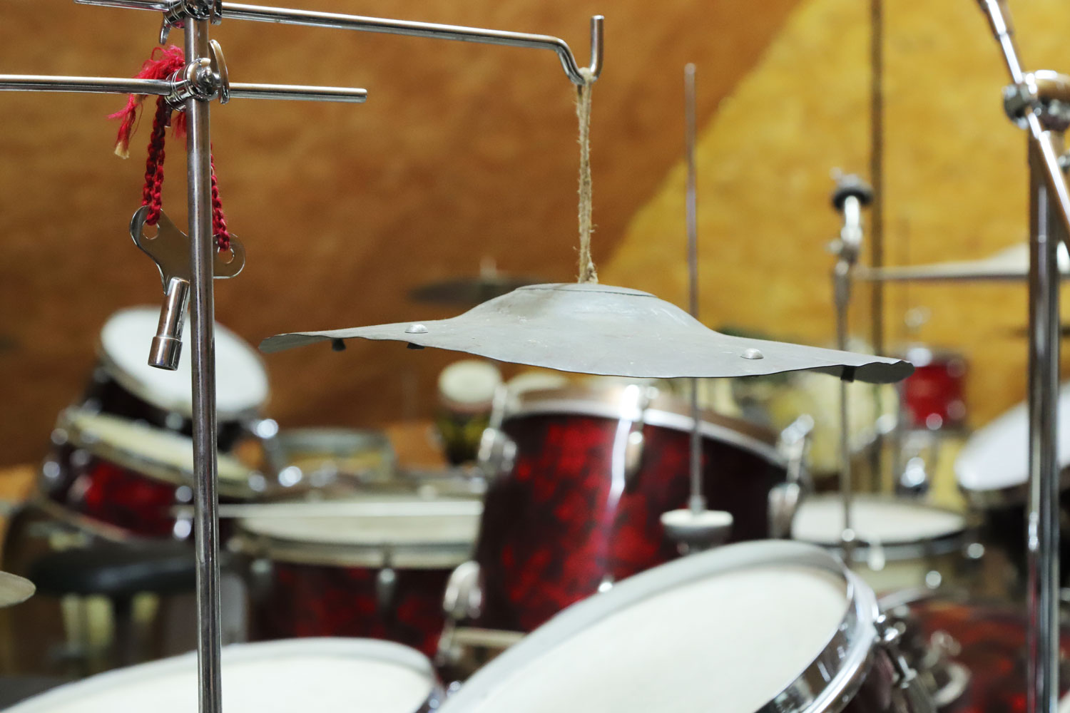 Ein scheinbar verbogenes, grau-metallisches Becken hängt an einem Strick von einer Halterung herunter. An dieser Hängt auch ein silberner Stimmschlüssel an einer roten Kordel. Im Hintergrund ist weiteres Schlagwerk zu erkennen.)