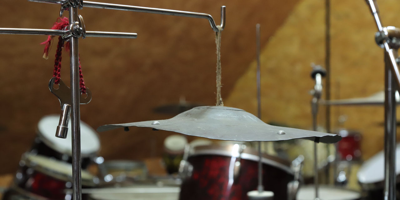 Ein scheinbar verbogenes, grau-metallisches Becken hängt an einem Strick von einer Halterung herunter. An dieser Hängt auch ein silberner Stimmschlüssel an einer roten Kordel. Im Hintergrund ist weiteres Schlagwerk zu erkennen.