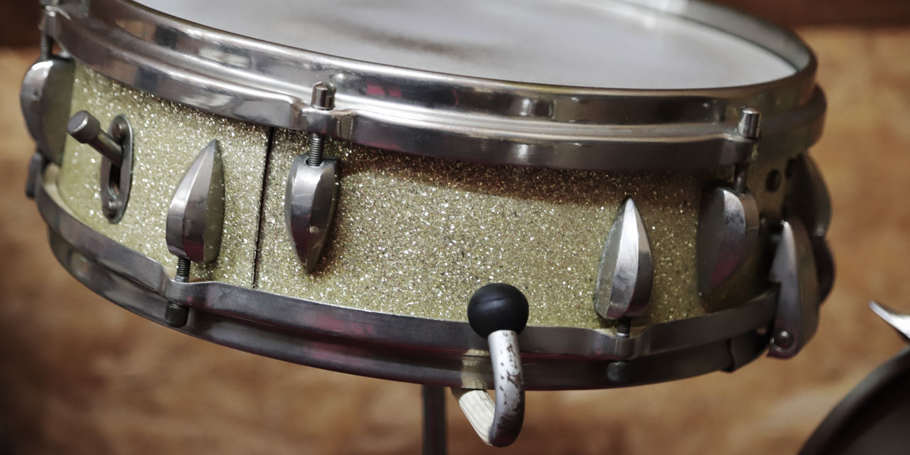 Eine Trommel mit Metallriemen und weißem Plastikfell, der Körper der Trommel besteht aus einem glitzernden Material, man kann einen metallenen Schalter an ihm erkennen. Unten an der Trommel ragt ein Metallhacken mit einer runden, schwarzen Gummikugel an der Spitze unter der Trommel hervor