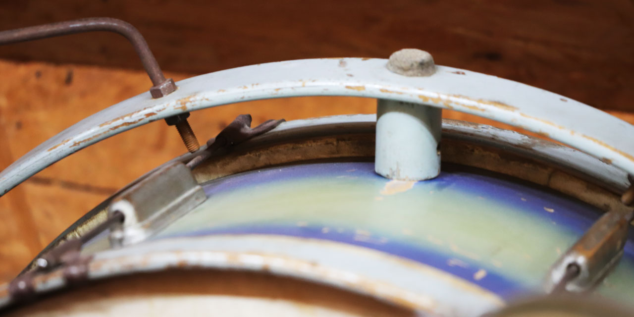 Man erkennt einen bläulich lackierten Metallriemen, der über einen Sockel an der Seite einer großen Trommel befestigt ist, welche man noch teilweise am unteren Bildrand sieht.