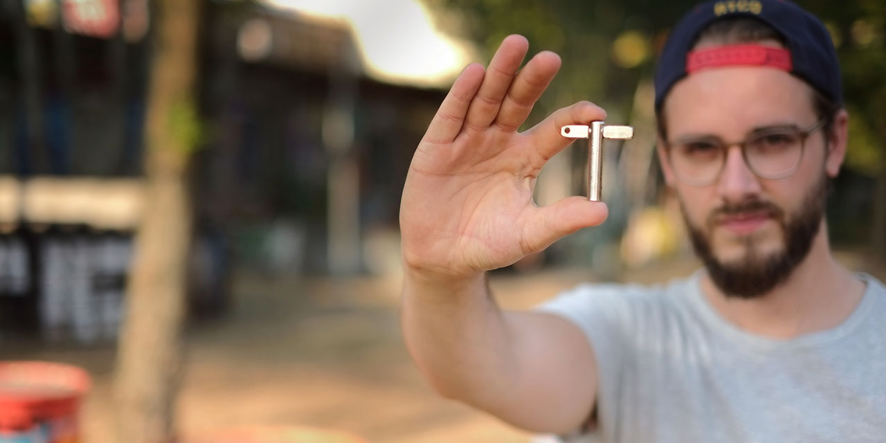 Ein Mann hält einen metallenen Stimmschlüssel zwischen Daumen und Zeigefinger der rechten Hand in die Kamera. Er hat braune Haare und Bart, trägt ein weißes T-Shirt und ein umgedrehtes Cap. Der Hintergrund ist verschwommen, man erkennt aber, dass das Foto im Freien geschossen wurde.