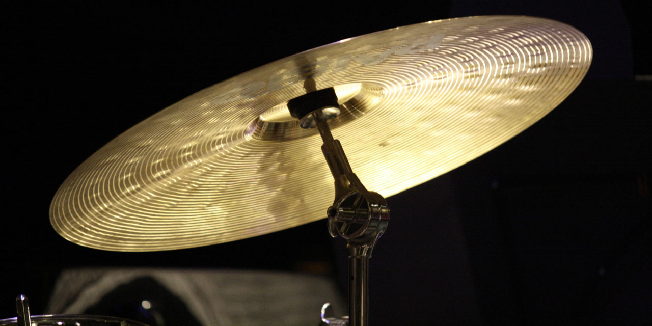 Ein gold-messingfarben glänzendes Becken auf einen dazugehörigen Ständer vor schwarzem Hintergrund