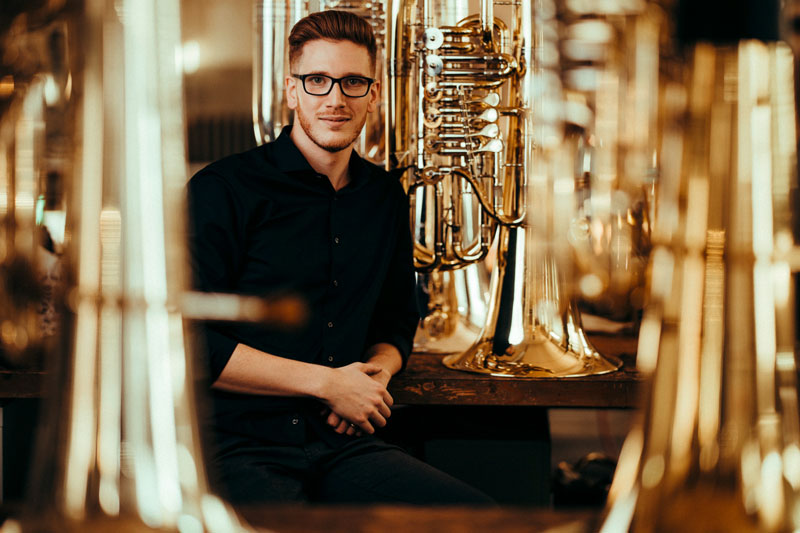 Constantin Hartwig stützt seinen Ellbogen auf einen Tisch, auf dem seine Tuba steht und lächelt in die Kamera: Um ihn herum stehen weitere Tuben auf Tischen. 