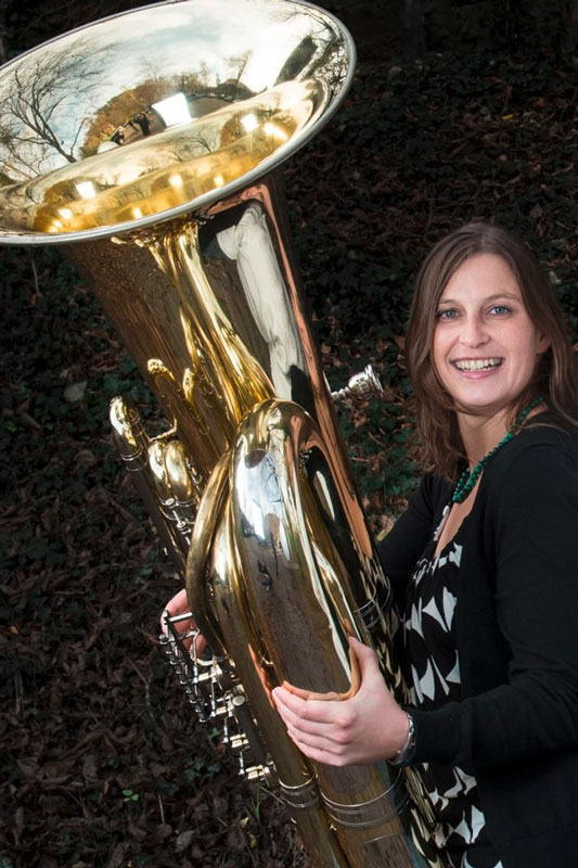 Corinna Dengler hält lächelnd ihre große Tuba neben sich in den Händen vor einem dunklen Hintergrund