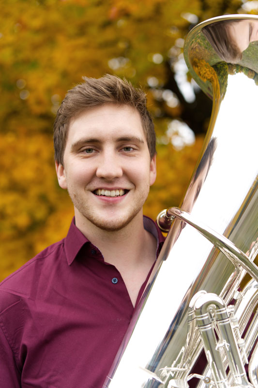 Donath Rehm im dunkelroten Hemd lächelt mit seiner Tuba in der Hand in die Kamera und im Hintergrund sind die Blätter eines Baumes zu sehen.