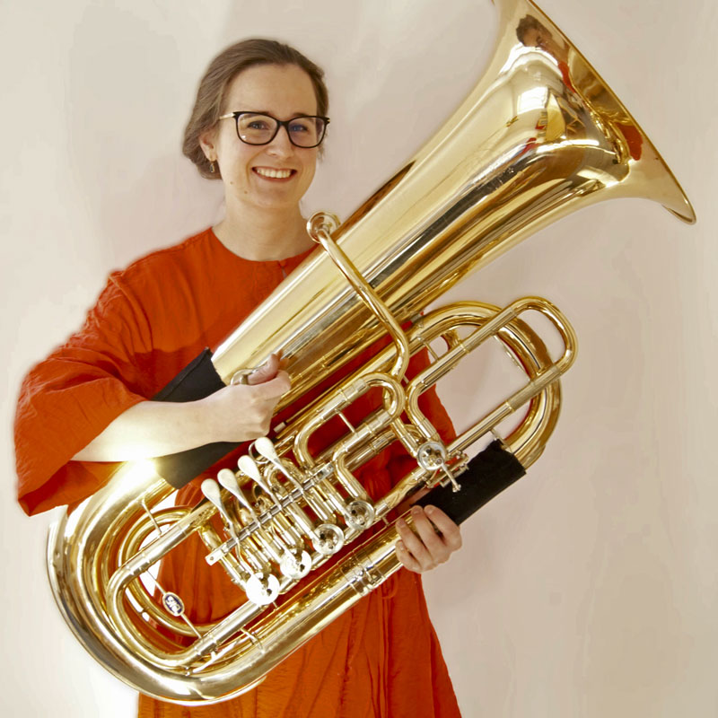 Henriette Buchholz steht lächelnd in einem roten Kleid mit ihrer Tuba in der Hand vor einem hellen Hintergrund.