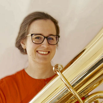 Henriette Bruchholz, blondbraune Haare hinten zusammengesteckt, orangenes Oberteil und schwarze Brille, hält Tuba vor sich und lächelt in Kamera 