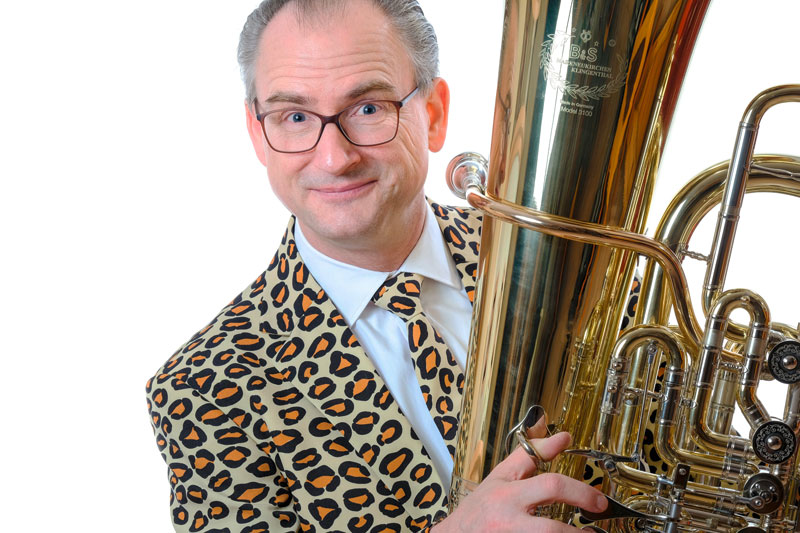 Jörg Wachsmuth hält in seinem Leopardenmuster Anzug seine Tuba in der Hand und lächelt in die Kamera vor einem weißen Hintergrund