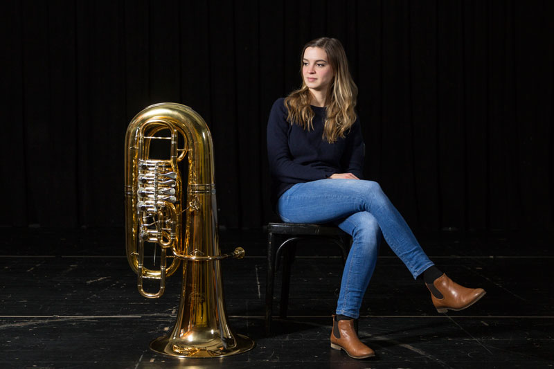 Ruth Ellendorff sitzt vor einem schwarzen Hintergrund auf einem Hocker, das eine Bein über das andere geschlagen, und schaut auf ihre Tuba, welche neben ihr auf dem Boden steht.