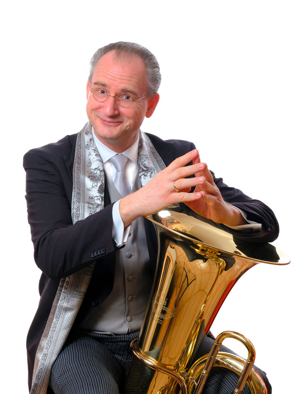 Der Schirmherr des IdJ 2024: Prof. Jörg Wachsmuth, kurze grauweiße Haare, Brille mit runden Gläsern, er stützt seine Arme auf den Trichter einer Tuba und trägt ein weißes Hemd samt weißer Krawatte, sowie ein langes schwarzes Jackett. 