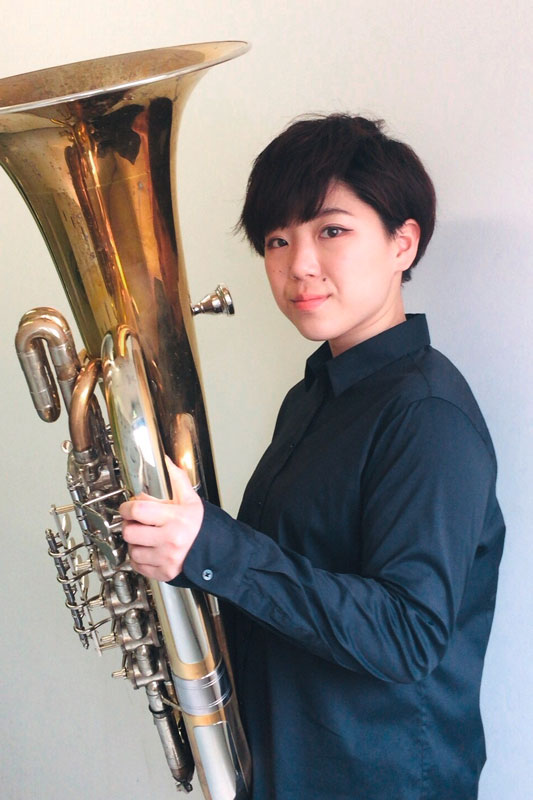 Shiho Murano steht in einem dunkelblauen Hemd mit ihrer Tuba in den Händen vor einem weißen Hintergrund und lächelt in die Kamera.