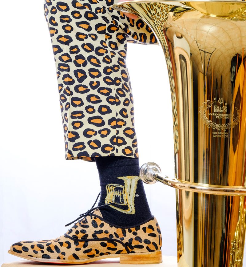 Der obere Teil einer (Trichter und Mundstück) sind neben einem 90° angewinkelten Bein zu sehen, dieses ist in eine Hose mit Leopardenmuster gekleidet, auch die Schuhe haben ein solches gelb-schwarzes Muster, die Socken mit goldener Tuba darauf sind zu sehen