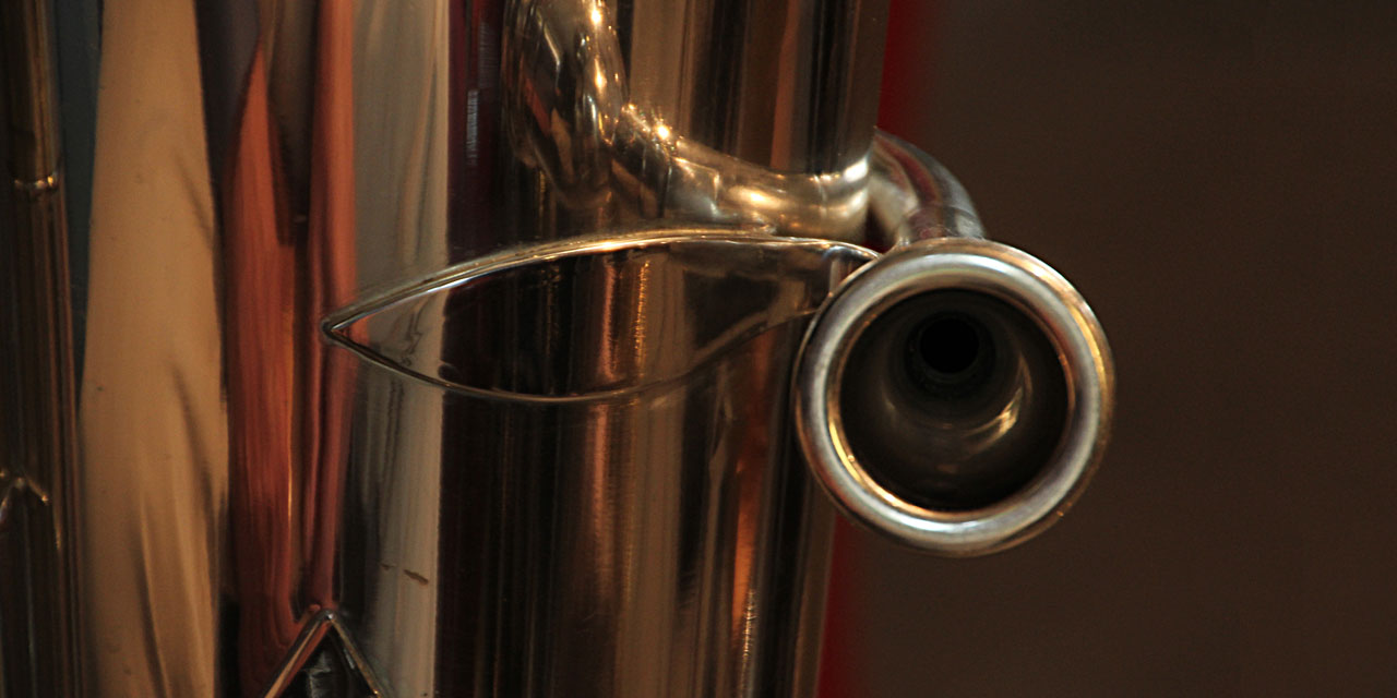 Ein goldglänzendes Tuba Mundstück mit dunkler Öffnung ist groß zu sehen. Es spiegelt sich im Rest der Tuba.