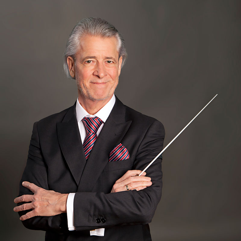 Walter Hilgers steht im schwarzen Anzug mit verschränkten Armen und einem Taktstock in der Hand vor einem dunkelgrauen Hintergrund und lächelt leicht in die Kamera.