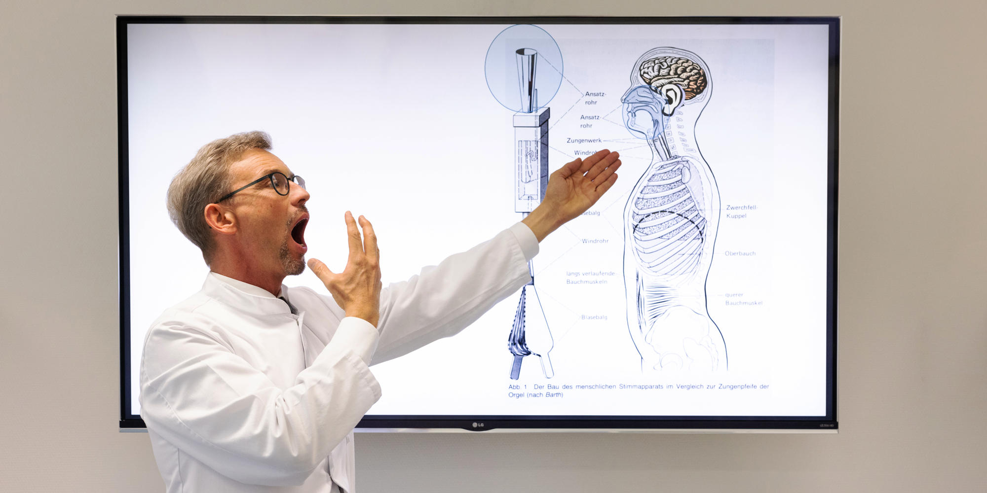 Der Schirmherr für Sachsen Professor Doktor Michael Fuchs präsentiert mit offenem Mund Ein Modell an der Wand, auf dem man die Funktion der Stimme erklären kann.