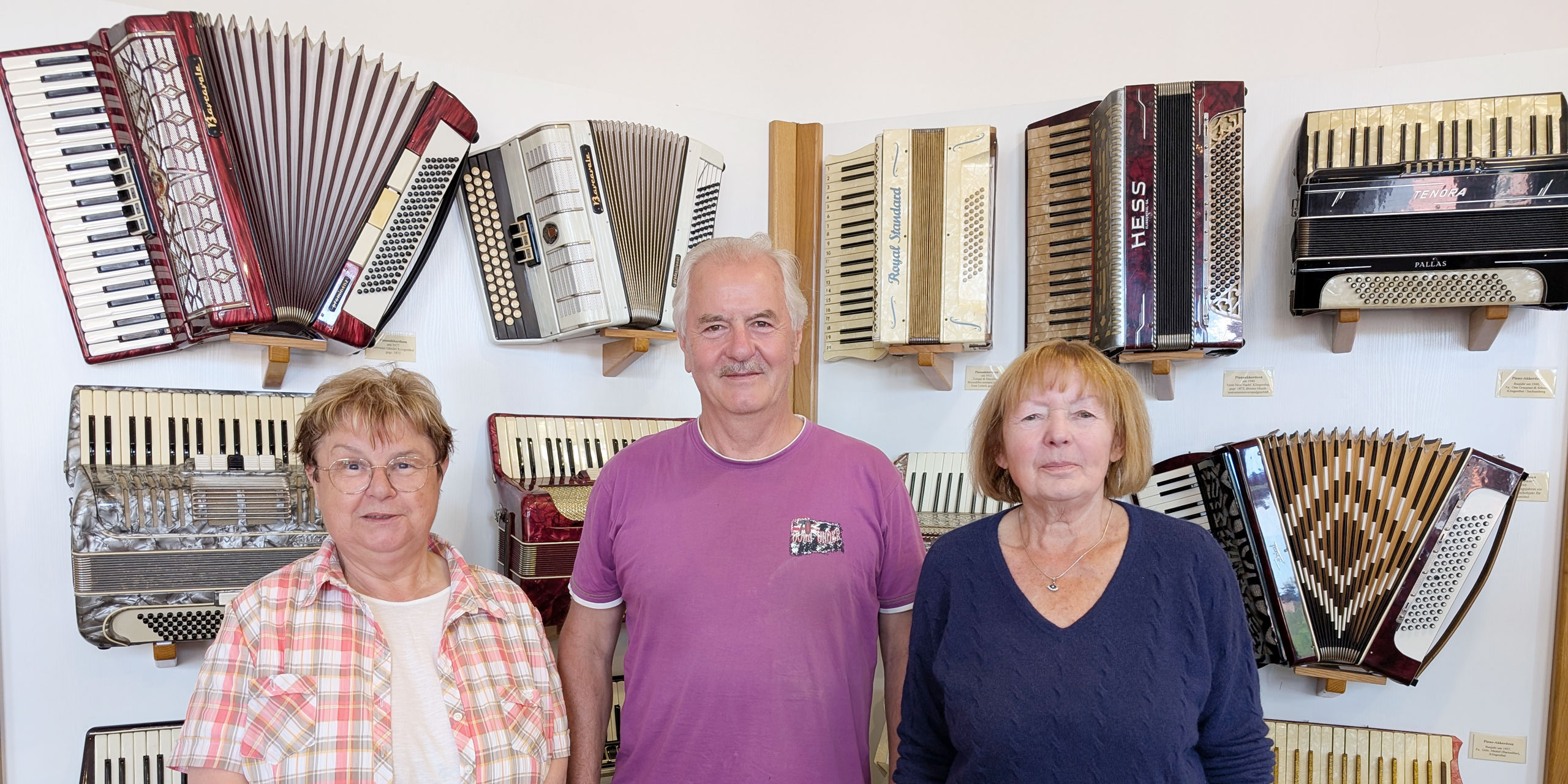 Drei Personen, zwei Damen und ein Herr in der Mitte, stehen vor einer Wand, an der zahlreiche Akkordeons hängend ausgestellt sind.