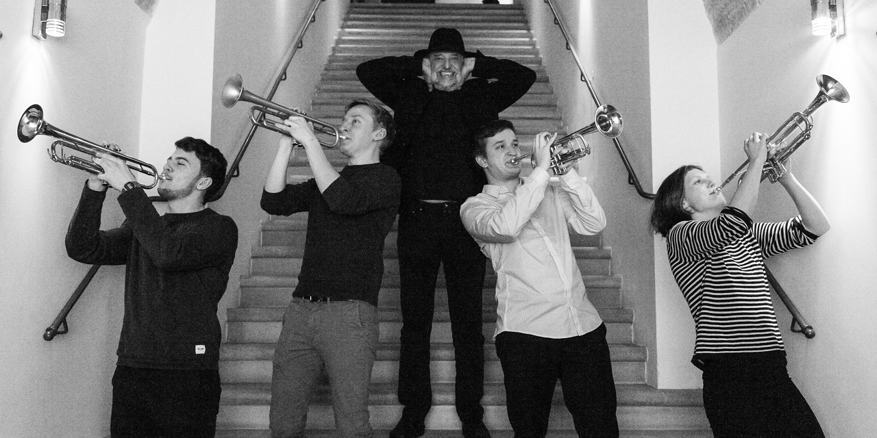 Vier Trompeten spielende Personen stehen nebeneinander vorne. Die zwei auf der rechten Seite heben ihr Instrument schräg nach oben in die rechte Bildrichtung und die zwei auf der anderen Seite halten ebenfalls ihre Instrumente in die entgegengesetzte Bildrichtung nach oben. In der Mitte hinter den vier Personen steht ein wenig erhöht auf einer Treppe ein älterer Mann mit einem schwarzen Hut, welcher sich die Ohren zuhält.