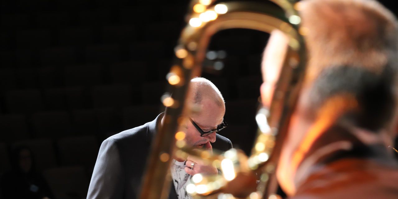 Im Vordergrund steht ein Posaunist und im Hintergrund der Dirigent fokussiert.