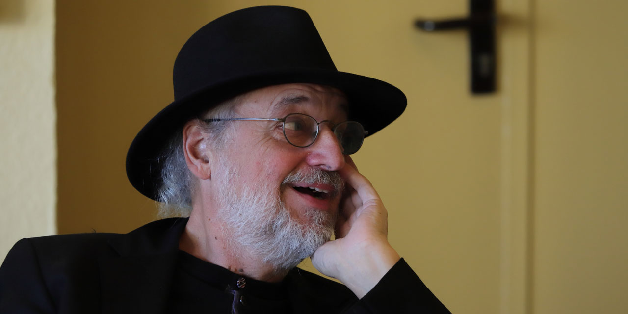 Eine Nahaufnahme eines älteren Mannes mit weißen Haaren und Bart, welcher eine Brille und einen schwarzen Hut trägt und lachend zur Seite schaut 