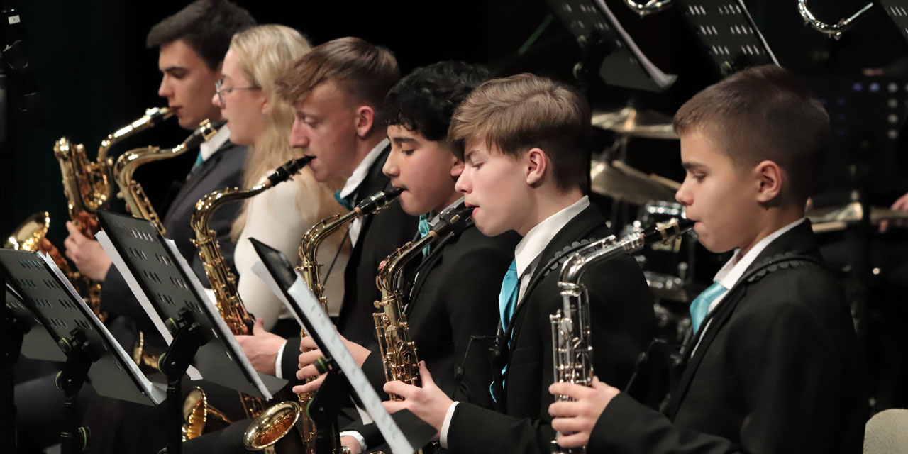 Es sind insgesamt sechs Saxophonspieler/-innen auf einer Bühne zu sehen. Sie tragen weiße, blaue und schwarze Konzertkleidung.